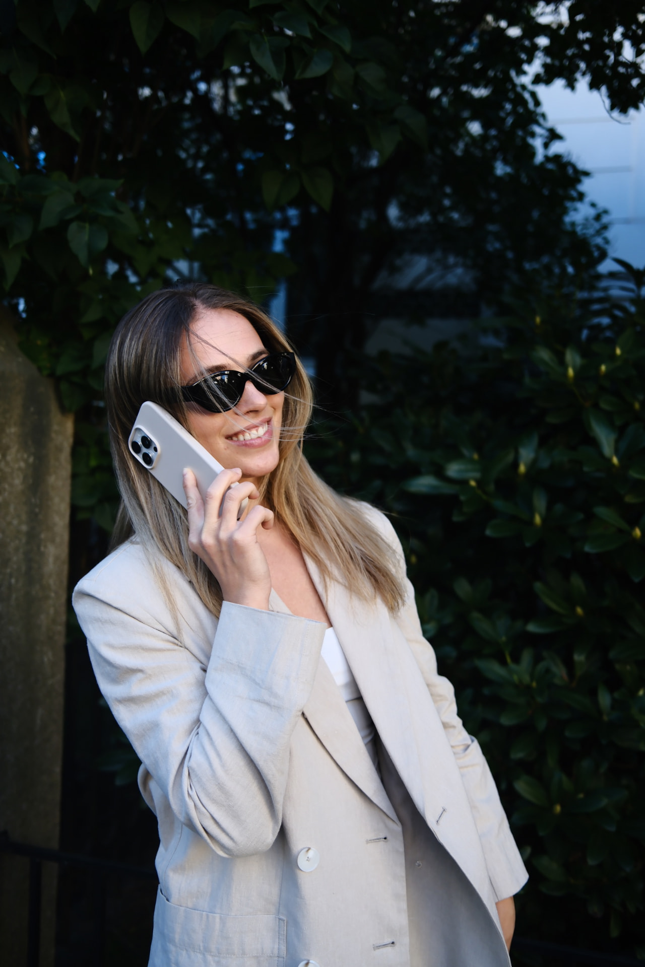 Blond kvinne i beige blazer med solbriller i telefonmotælling mot mørk grønn bakgrunn — KRE@IVE STUDIOS editorial